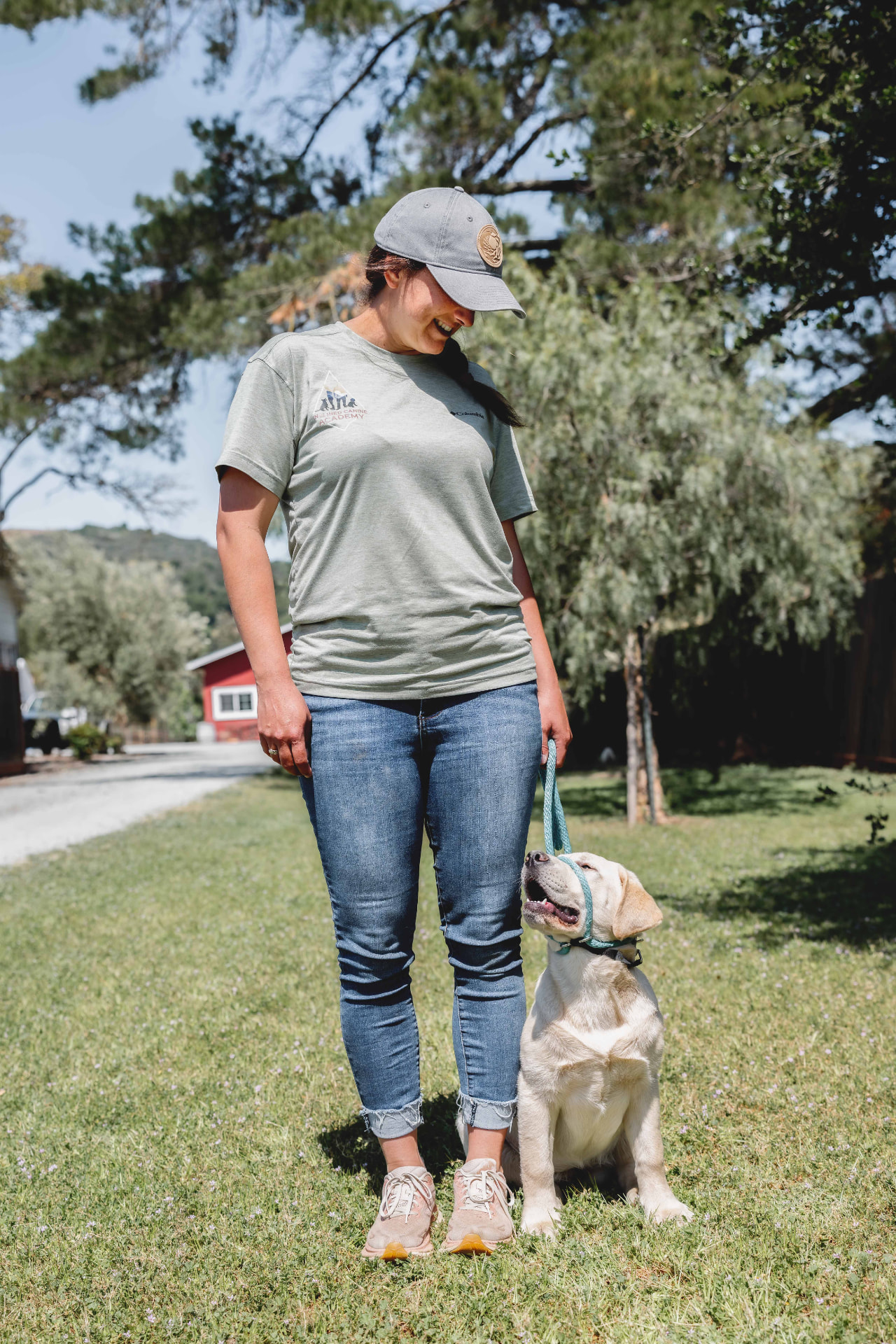 A woman in an Inclined Canine Academy t-shirt smiling down at a yellow Labrador puppy sitting on the grass at the Morgan Hill facility